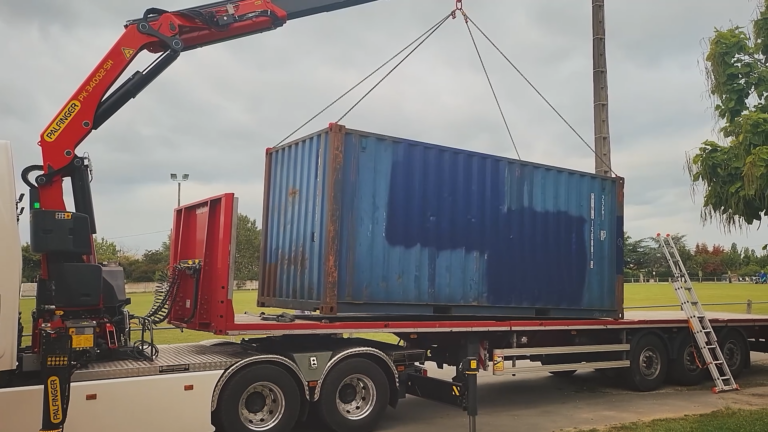 Grue soulevant un container bleu de 20 pieds d'occasion près d'un stade.