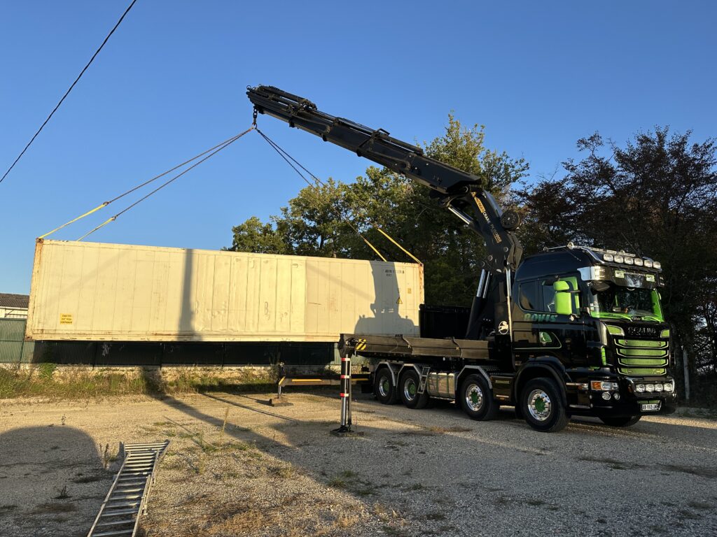 Un camion équipé d'une grue soulevant un grand conteneur isotherme dans un espace extérieur.