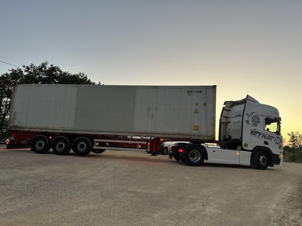 Un camion blanc portant le logo "Veynat 16" transporte un grand conteneur isotherme blanc, garé sur un terrain gravillonné au crépuscule.