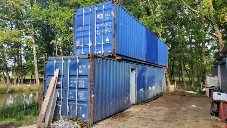 Deux grands conteneurs bleus empilés l'un sur l'autre à l'extérieur, à côté d'un bâtiment et devant des arbres, avec divers objets et matériaux autour.