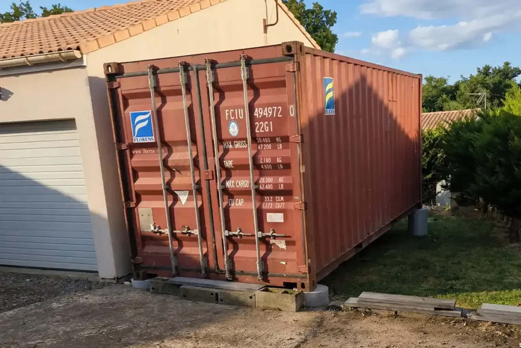 Container maritime 20 pieds d'occasion brun-rouge posé en abri de jardin sur plots béton contre un garage à Aigonnay (79)