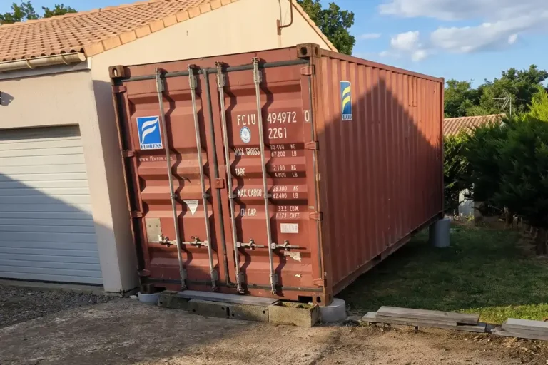 Container maritime 20 pieds d'occasion brun-rouge posé en abri de jardin sur plots béton contre un garage à Aigonnay (79)
