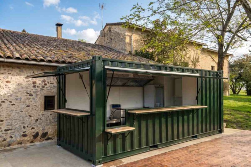 Container snack vert sapin adossé à un bâtiment en pierre, avec comptoir bois et terrasse, au Domaine Laborde à Paulhiac