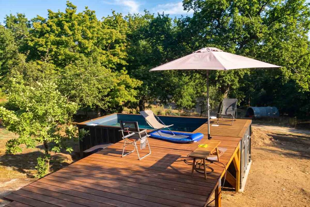 Terrasse en bois au niveau des margelles d'une piscine container, avec parasol et chaises, à Abjat-sur-Bandiat